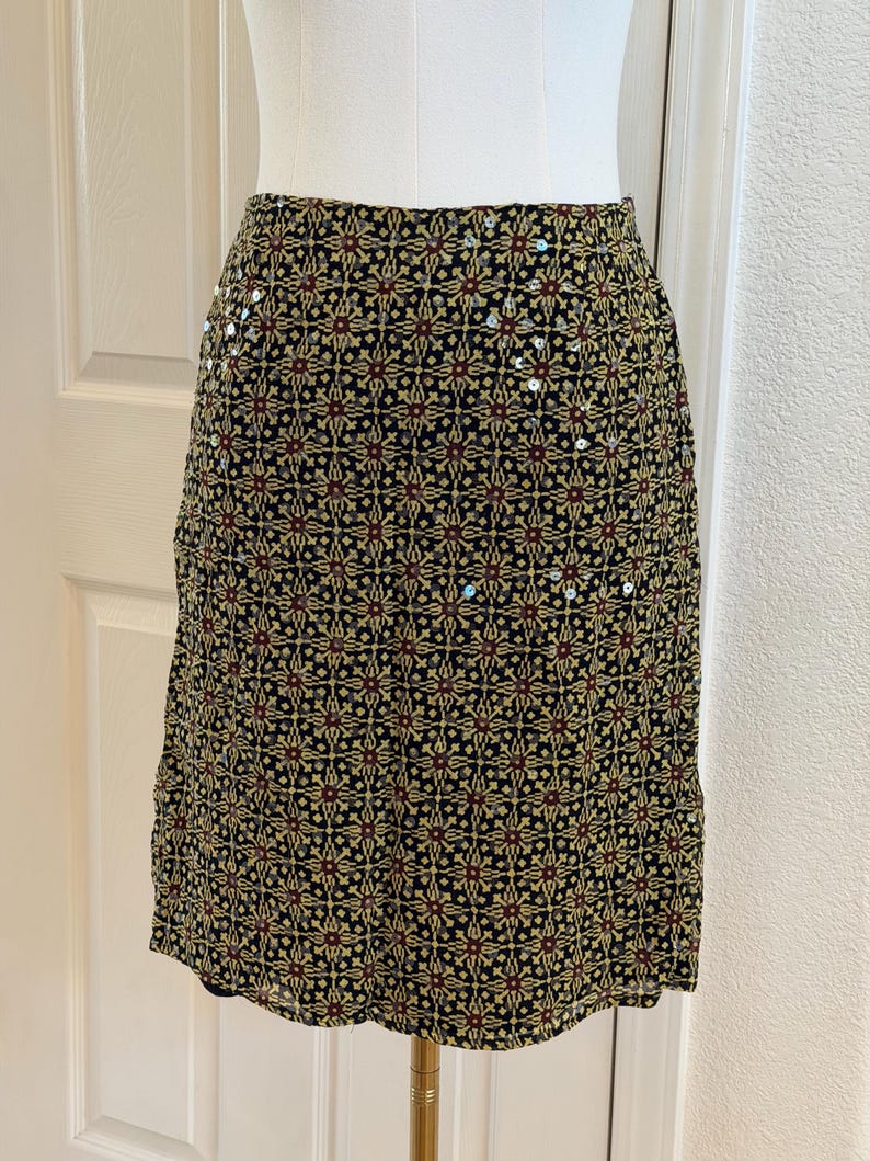 May include: A knee-length skirt featuring a black base with a repeating geometric pattern in shades of yellow, red, and green. The fabric is embellished with small, scattered silver sequins that catch the light across the surface of the garment.