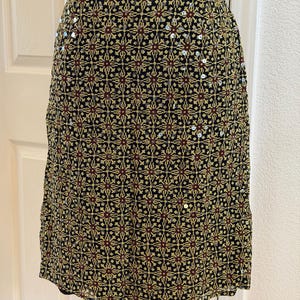 May include: A knee-length skirt featuring a black base with a repeating geometric pattern in yellow and dark red. The fabric is embellished with scattered, small, reflective silver sequins throughout the design, creating a subtle shimmer across the surface.