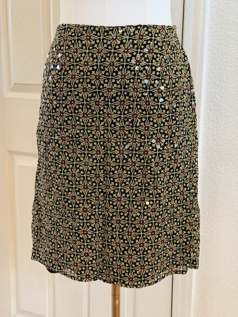 May include: A knee-length skirt featuring a black base with a repeating geometric pattern in yellow and dark red. The fabric is embellished with scattered, small, reflective silver sequins throughout the design, creating a subtle shimmer across the surface.