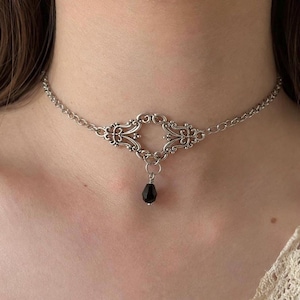 Könnte beinhalten: Eine silberfarbene Metall-Halskette als Choker mit einem zentralen, filigranen Dekorelement mit symmetrischen Schnörkelmustern. Eine einzelne kleine, facettierte schwarze tropfenförmige Perle hängt in der Mitte des Metallstücks.