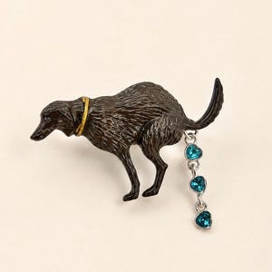 May include: A dark brown dog-shaped brooch with a gold-tone collar. A silver-tone chain hangs from the dog's rear, featuring three small, dangling, heart-shaped blue crystals. The dog is depicted in a crouching posture with its tail raised.