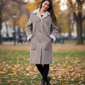 Może przedstawiać: Długi, jasnobrązowy płaszcz z kremowym kołnierzem i mankietami z owczej skóry. Płaszcz ma duże kieszenie i zapięcia na guziki. Osoba nosi czarne legginsy i brązowe buty. Tło to park z jesiennymi liśćmi.