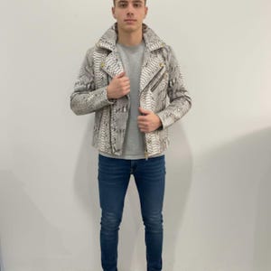 Może przedstawiać: Szara i biała skórzana kurtka ze wzorem skóry węża, z kołnierzem z klapami i asymetrycznym zapięciem na zamek błyskawiczny. Kurtka ma złote okucia i jest połączona z szarym t-shirtem, niebieskimi dżinsami i białymi trampkami. Stylowa odzież wierzchnia.