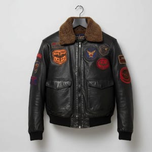 May include: Black leather bomber jacket with a brown shearling collar. The jacket features multiple colorful patches on the chest and sleeves, two front pockets, and a black ribbed hem and cuffs. The jacket is hanging on a black hanger.