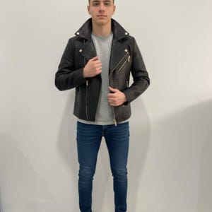 Może przedstawiać: Czarna, teksturowana skórzana kurtka z kołnierzem z klapami i złotymi zamkami błyskawicznymi. Kurtka jest połączona z szarą koszulką, niebieskimi dżinsami i białymi trampkami. Stylowa odzież wierzchnia.