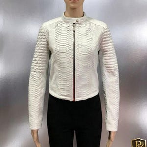 May include: A white, textured jacket with a snakeskin pattern. The jacket features a stand-up collar, a full-length zipper, and long sleeves. The jacket is displayed on a mannequin, paired with black pants.