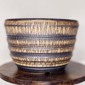 May include: A cylindrical ceramic planter featuring a textured exterior with alternating horizontal bands of dark brown and light tan. The surface displays a vertical, incised grid pattern throughout, with a smooth, dark brown base and a wide, open top.