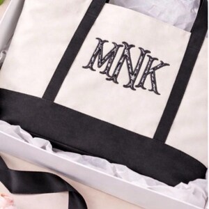 May include: A cream-colored canvas tote bag with black fabric handles and a black base. The bag features the embroidered black letters MNK in a decorative serif font. It is presented inside a white gift box with white tissue paper.