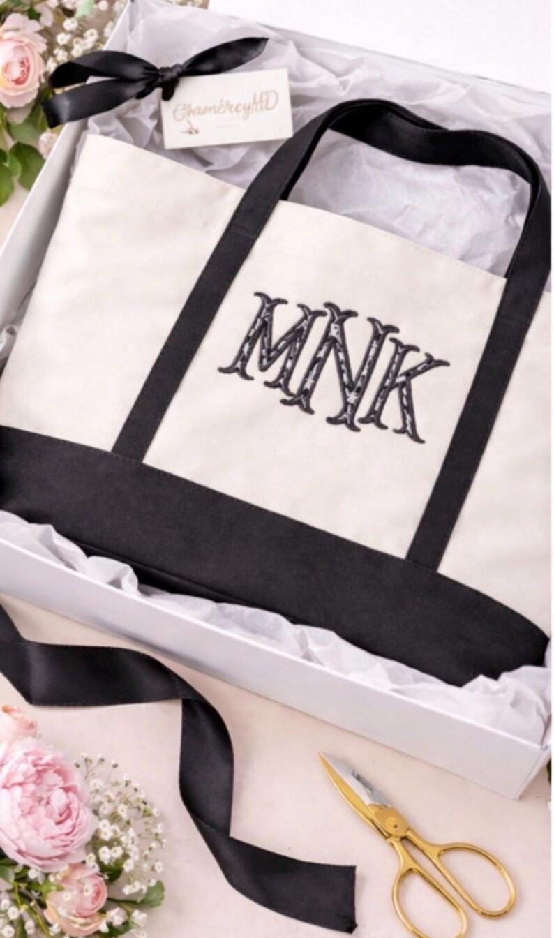 May include: A cream-colored canvas tote bag with black fabric handles and a black base. The bag features the embroidered black letters MNK in a decorative serif font. It is presented inside a white gift box with white tissue paper.