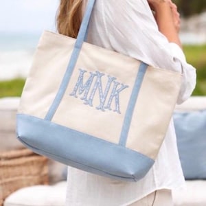 May include: A cream-colored canvas tote bag featuring a light blue base, matching light blue shoulder straps, and the embroidered monogram letters MNK in a light blue decorative font centered on the front panel of the bag.