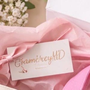 May include: A white gift box with pink tissue paper and a pink ribbon. A small white card with gold lettering that reads "Cham&egrave;rcyHlD" is placed inside the box. The overall aesthetic is soft and feminine.
