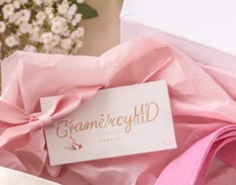 May include: A white gift box with pink tissue paper and a pink ribbon. A small white card with gold lettering that reads "Cham&egrave;rcyHlD" is placed inside the box. The overall aesthetic is soft and feminine.