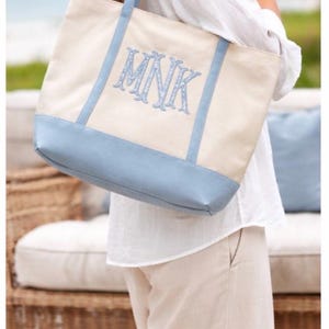 May include: Cream-colored canvas tote bag with light blue accents and handles. The bottom and the handles are light blue. The bag features the embroidered initials "MNK" in a light blue script. A stylish and practical accessory.