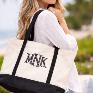 May include: Cream-colored tote bag with black accents and handles. The bag features the embroidered initials "MNK" in a decorative font. The bottom of the bag is black. The bag is carried over the shoulder.