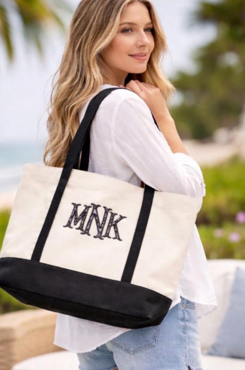 May include: Cream-colored tote bag with black accents and handles. The bag features the embroidered initials "MNK" in a decorative font. The bottom of the bag is black. The bag is carried over the shoulder.