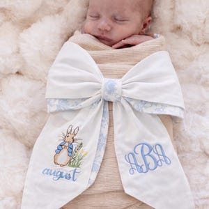 May include: A newborn baby wrapped in a beige blanket, adorned with a large white bow featuring a blue floral pattern. The bow has an embroidered Peter Rabbit design and the name "august" in blue script, along with a monogram.