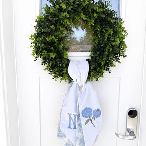May include: A circular green eucalyptus wreath featuring a white fabric ribbon tied at the bottom. The ribbon displays a blue letter N and a blue hydrangea flower illustration on a white background with a blue and white toile pattern on the reverse.