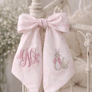May include: A pink fabric bow with a floral pattern is tied to a white bed frame. The bow has two hanging panels, one with embroidered initials and the other with a Peter Rabbit design.