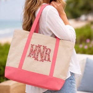 May include: A beige canvas tote bag with bright pink trim and handles. The bag features a large, embroidered monogram with the letters MNR in a pink and leopard print pattern, centered on the front panel of the bag above the pink base.