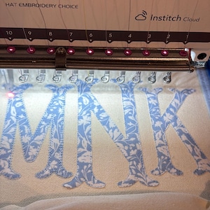 May include: An embroidery machine is shown in operation, stitching the letters "MNK" onto a light-colored fabric. The letters are in a light blue floral pattern. The machine has the "BAI" logo and "Institch Cloud" text.