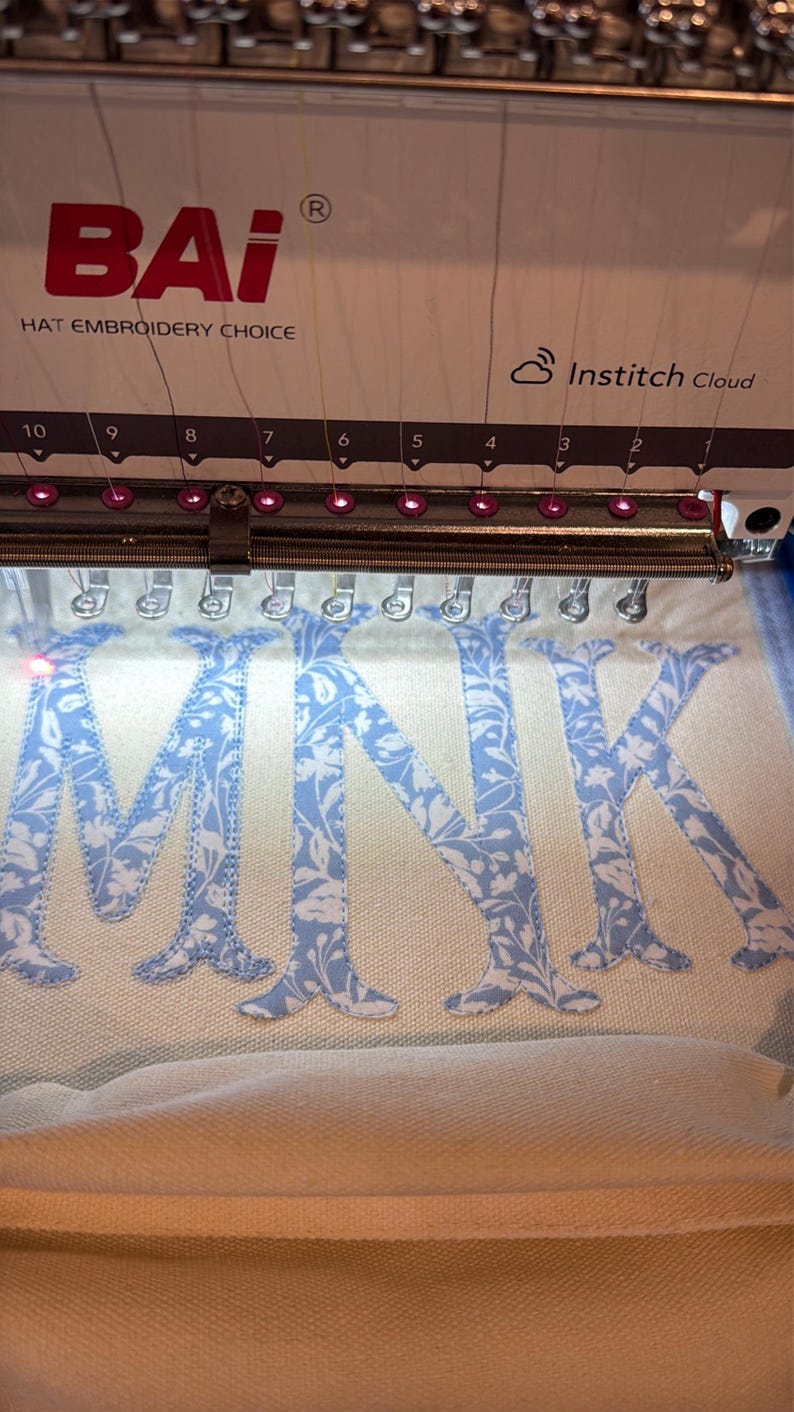 May include: An embroidery machine is shown in operation, stitching the letters "MNK" onto a light-colored fabric. The letters are in a light blue floral pattern. The machine has the "BAI" logo and "Institch Cloud" text.