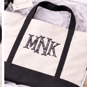 May include: A cream-colored canvas tote bag with a black base and black handles. The front of the bag features the embroidered letters MNK in a dark, decorative font. The bag is presented inside a white box with white tissue paper.