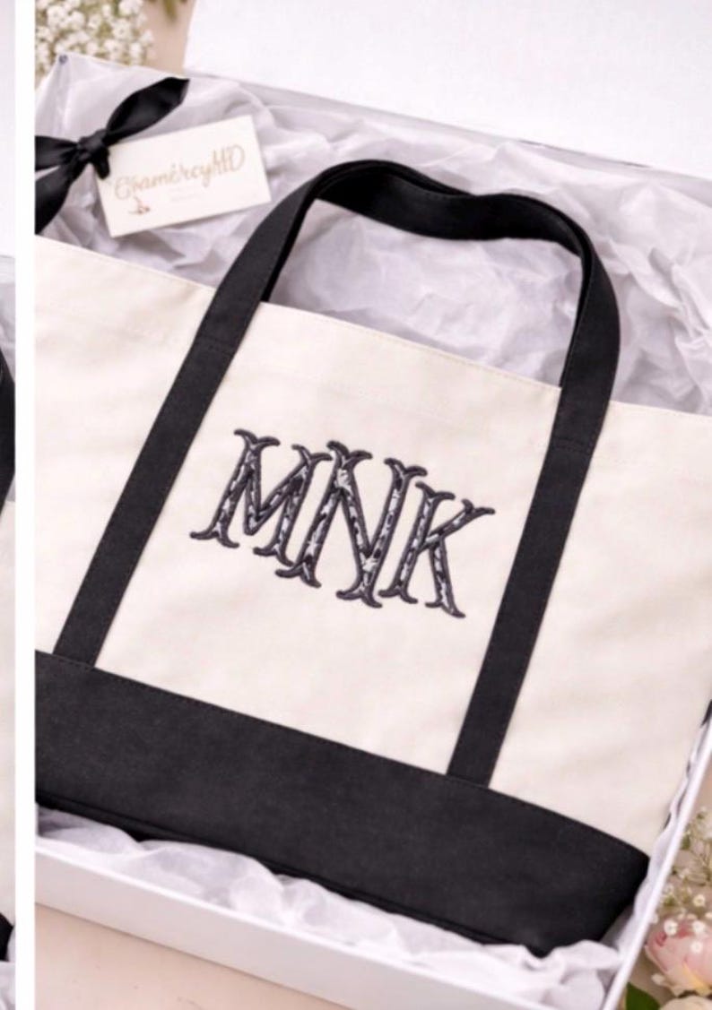 May include: A cream-colored canvas tote bag with a black base and black handles. The front of the bag features the embroidered letters MNK in a dark, decorative font. The bag is presented inside a white box with white tissue paper.