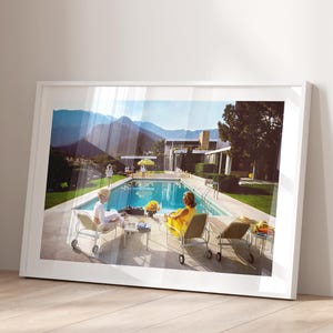 Könnte beinhalten: Ein gerahmter Druck einer Vintage-Szene mit einem Swimmingpool, Liegestühlen und Personen. Das Bild hat eine Retro-Ästhetik mit einem Haus im Stil der Moderne der Jahrhundertmitte im Hintergrund. Das Poolwasser ist leuchtend blau und der Himmel ist klar.