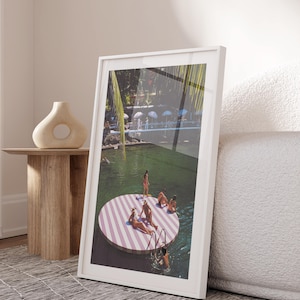 May include: A framed art print showing a pool scene with people on a striped circular platform. The platform has pink and white stripes. The print is in a white frame and is leaning against a white sofa. The scene is set in a sunny location.