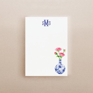 May include: A white rectangular notepad featuring a dark blue monogram with the letters SMB at the top center and a watercolor illustration of pink flowers in a blue and white patterned vase positioned in the bottom right corner.