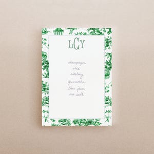 May include: A rectangular notepad with a white center and a green border featuring a detailed toile pattern. The top center displays the monogram LCY in green. Below, the handwritten text reads: champagne, rosé, riesling, grenadine, lime juice, sea salt.