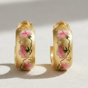 May include: A pair of gold-colored hoop earrings. Each earring features a floral design with pink flowers and green stems. The earrings also have star-shaped accents and small, clear gemstones.