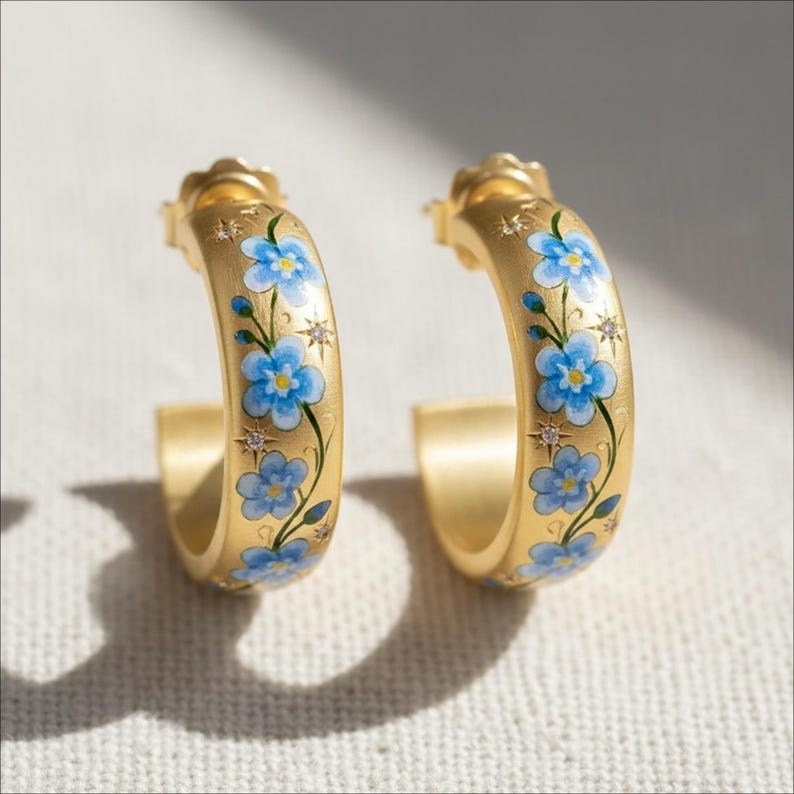 May include: A pair of gold-colored hoop earrings. Each earring is adorned with a delicate floral design featuring blue flowers and green stems. Small star-shaped accents and tiny crystals add sparkle to the design.