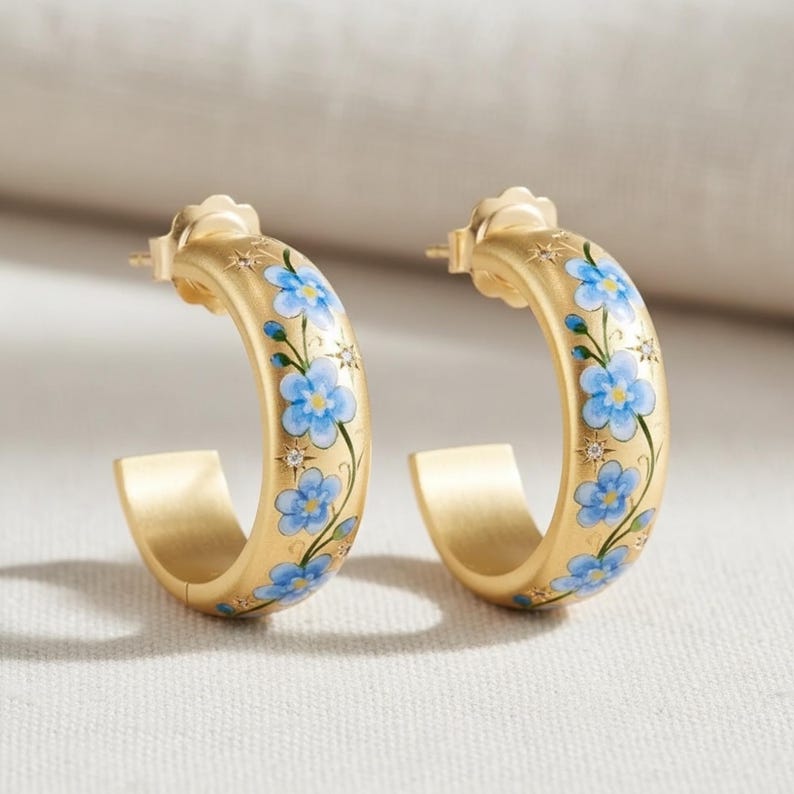 May include: A pair of gold-tone hoop earrings. Each earring is decorated with blue floral designs and small, sparkling accents. The earrings have a matte finish and a classic hoop shape, perfect for everyday wear.