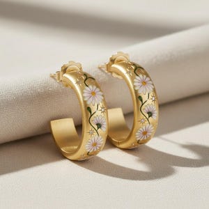 May include: A pair of gold-colored hoop earrings. Each earring is decorated with white and pink daisy flowers and green stems. The earrings have a matte finish and are displayed on a white surface.
