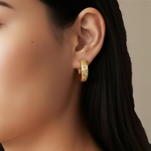 May include: Gold-colored hoop earrings with a floral pattern. The earrings are worn on the ear and are visible in a close-up shot. The earrings have a detailed, embossed design. The background is a neutral color, and the focus is on the jewelry.