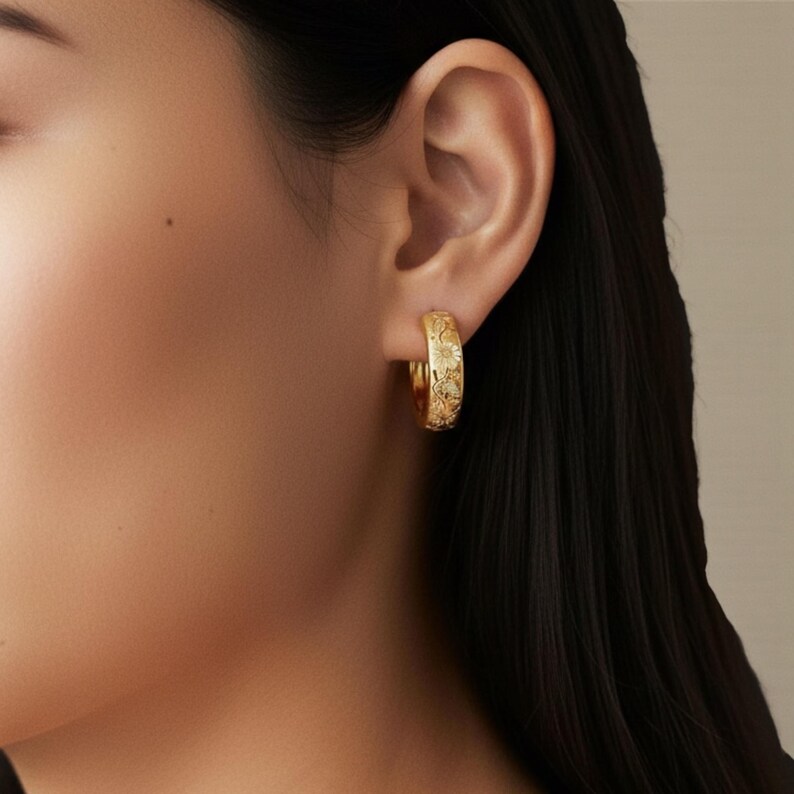 May include: Gold-colored hoop earrings with a floral pattern. The earrings are worn on the ear and are visible in a close-up shot. The earrings have a detailed, embossed design. The background is a neutral color, and the focus is on the jewelry.
