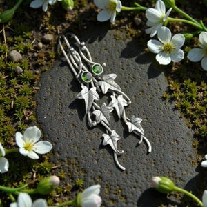 Puede incluir: Pendientes colgantes plateados con diseño de hojas y pequeñas gemas verdes. Los pendientes están colocados sobre una piedra gris oscura, rodeados de flores blancas y vegetación. Tema inspirado en la naturaleza.