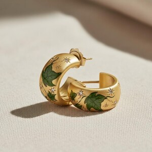 May include: Gold-tone hoop earrings with green leaf and star designs. The earrings have a matte finish and are adorned with small, sparkling crystals. The design features a botanical theme.