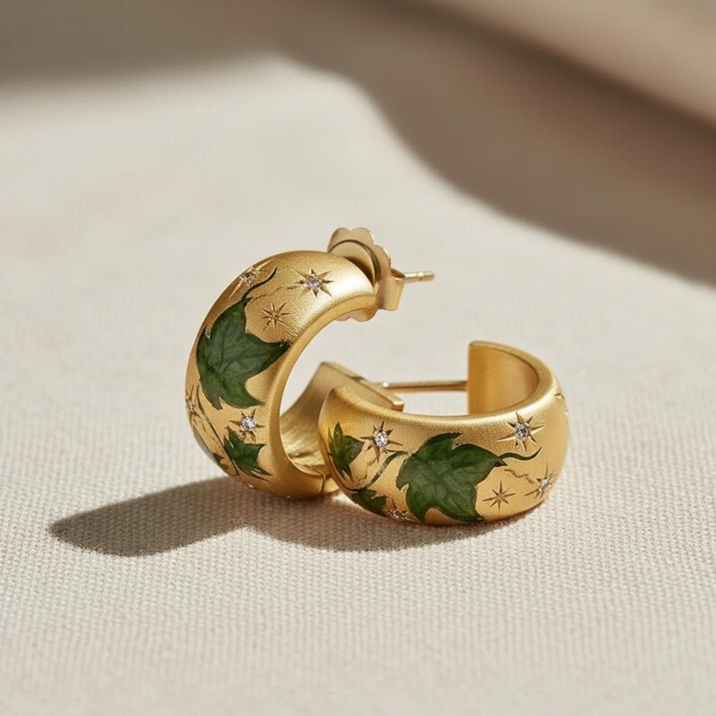 May include: Gold-tone hoop earrings with green leaf and star designs. The earrings have a matte finish and are adorned with small, sparkling crystals. The design features a botanical theme.