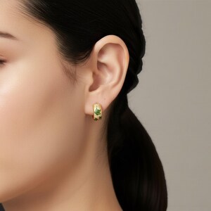 May include: Gold-toned hoop earrings with a green leaf design. The earrings are a small, curved shape, with a detailed green leaf pattern on the front. The earrings are worn on the ear.