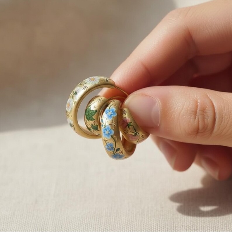 May include: Gold-toned hoop earrings with hand-painted floral designs. The earrings feature white daisies, blue forget-me-nots, and pink flowers with green leaves. The earrings are held in a hand.