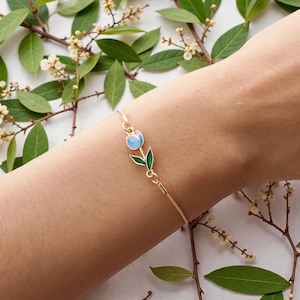 Può includere: Bracciale dorato con un design a tulipano. Il tulipano ha un fiore azzurro e foglie verdi. Il bracciale è delicato e ha un design a catena.