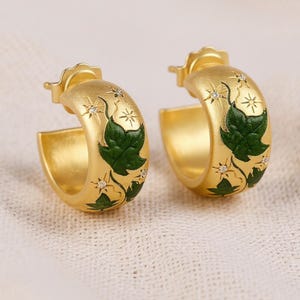 May include: A pair of gold-colored hoop earrings. Each earring features a green leaf design with small star-shaped accents and tiny clear stones. The earrings have a textured finish and a classic hoop shape.