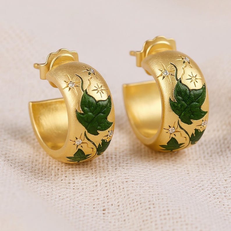 May include: A pair of gold-colored hoop earrings. Each earring features a green leaf design with small star-shaped accents and tiny clear stones. The earrings have a textured finish and a classic hoop shape.