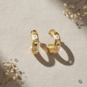 May include: A pair of gold-colored hoop earrings. Each earring features a floral design with white and lavender daisy-like flowers. The earrings are set against a neutral background with small decorative elements.