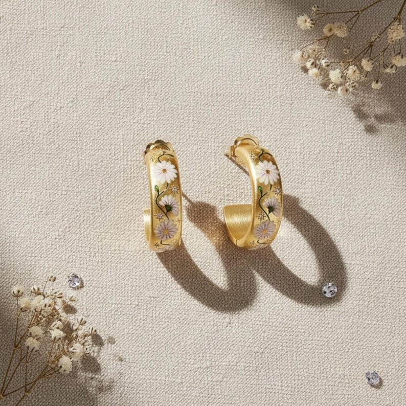 May include: A pair of gold-colored hoop earrings. Each earring features a floral design with white and lavender daisy-like flowers. The earrings are set against a neutral background with small decorative elements.