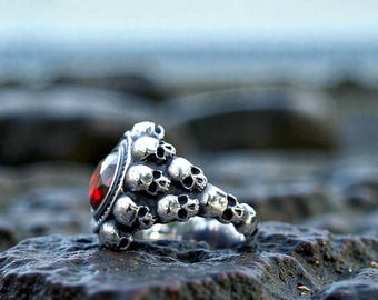 May include: A silver ring featuring a cluster of small skulls and a large, oval-shaped, red gemstone. The ring is set against a blurred background of a rocky surface and the ocean.