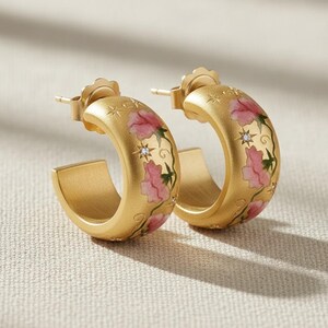 May include: A pair of gold-colored hoop earrings. Each earring features a floral design with pink flowers and green vines. Small star and diamond accents are also present. The earrings have a post-back closure.