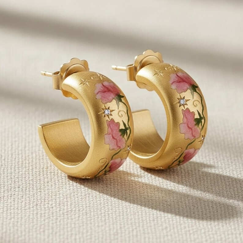 May include: A pair of gold-colored hoop earrings. Each earring features a floral design with pink flowers and green vines. Small star and diamond accents are also present. The earrings have a post-back closure.